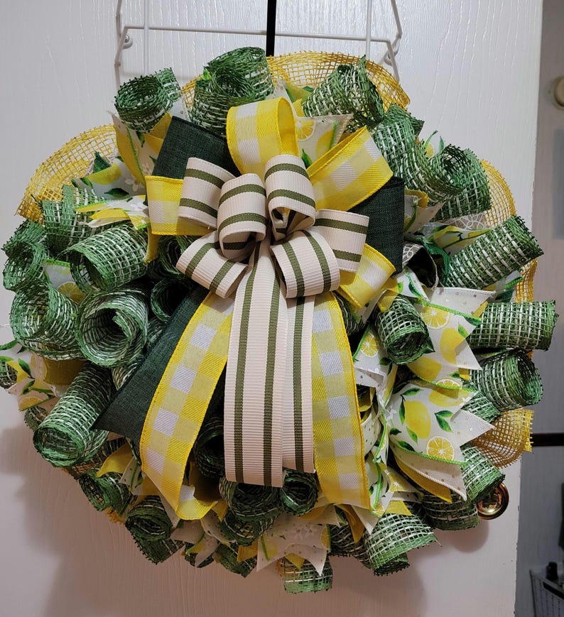 May include: A circular decorative wreath with a lemon theme. It features yellow, green, and white ribbons, with a large bow at the centre. The ribbons are arranged in a circular pattern, creating a full and textured appearance for a door or wall.