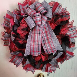 May include: A red, black, and gray plaid wreath with a large bow. The wreath is made of mesh and has a rustic look.