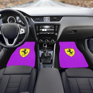Pu&ograve; includere: Tappetini per auto viola con il logo Ferrari giallo e nero. I tappetini sono all'interno di un'auto con cruscotto, volante e sedili neri. L'auto &egrave; parcheggiata su una strada.