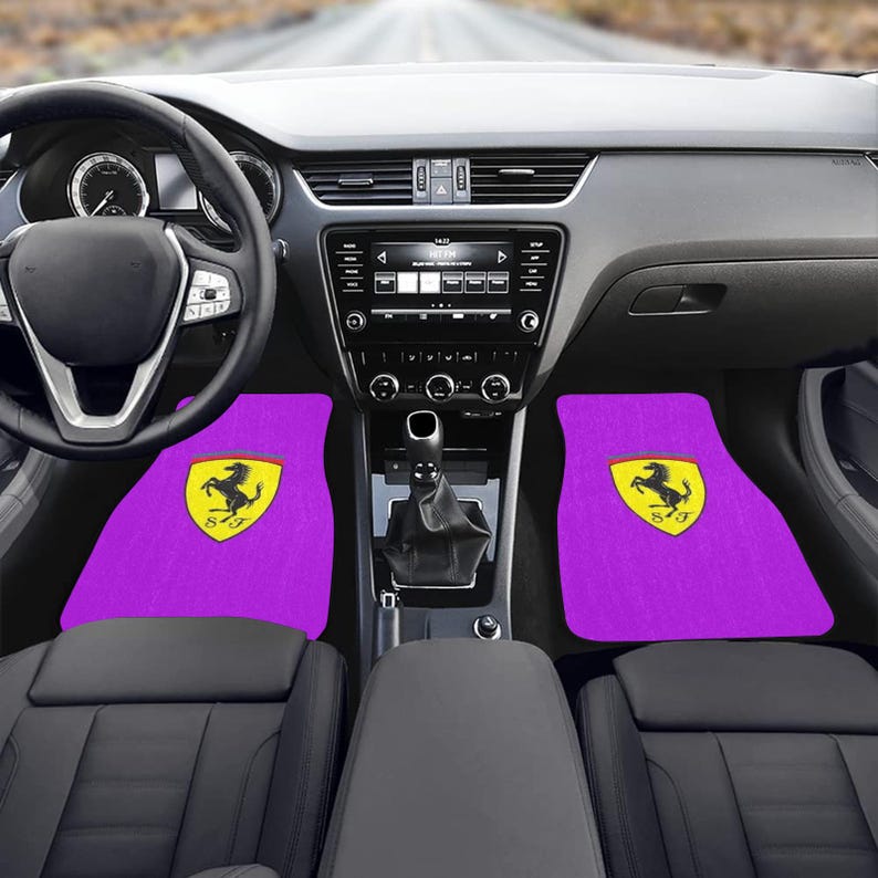 Pu&ograve; includere: Tappetini per auto viola con il logo Ferrari giallo e nero. I tappetini sono all'interno di un'auto con cruscotto, volante e sedili neri. L'auto &egrave; parcheggiata su una strada.