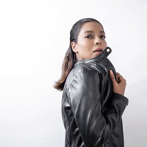 Peut inclure: Une femme portant une veste en cuir noire avec un col zipp&eacute;. Elle regarde par-dessus son &eacute;paule et a la main sur le col.