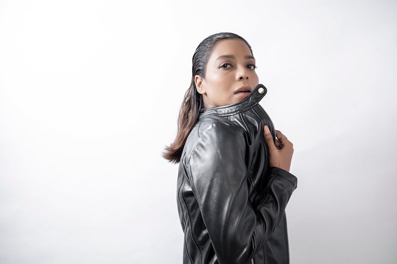 Peut inclure: Une femme portant une veste en cuir noire avec un col zipp&eacute;. Elle regarde par-dessus son &eacute;paule et a la main sur le col.