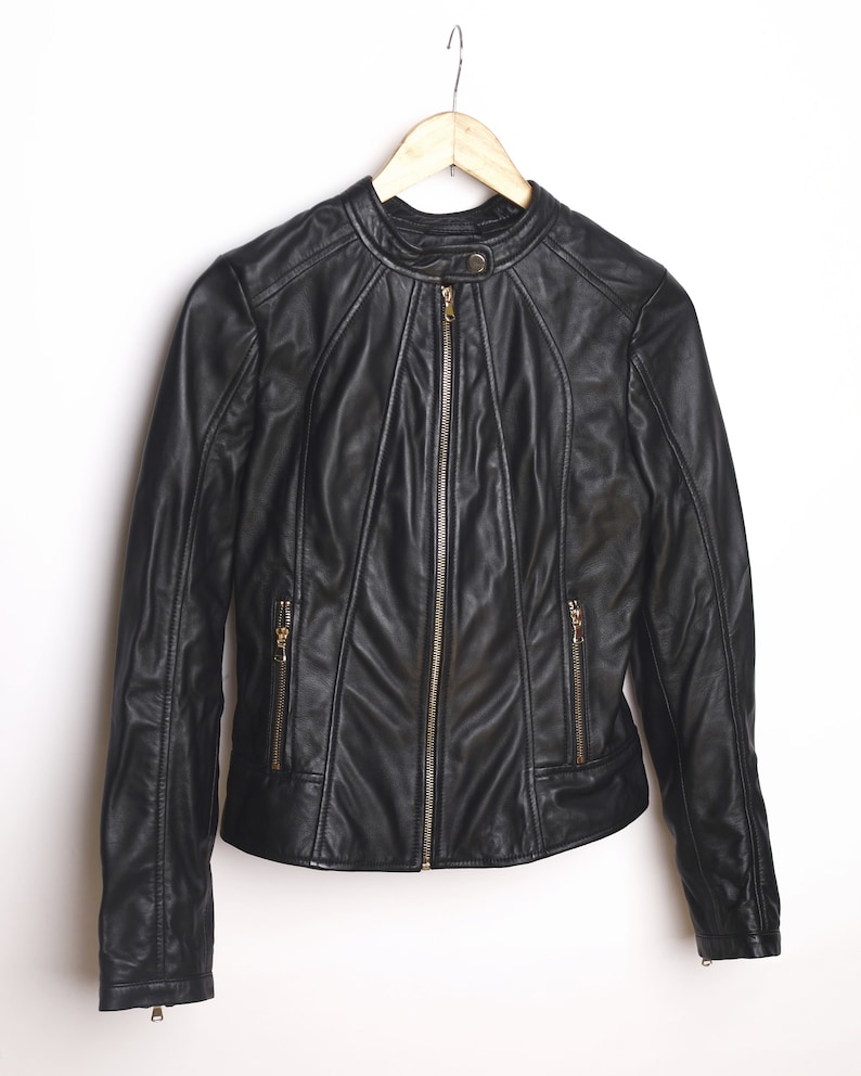 Peut inclure: Une veste en cuir noire avec une fermeture &eacute;clair et des poches dor&eacute;es. La veste est suspendue &agrave; un cintre en bois.