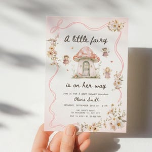 May include: A baby shower invitation with a pink border and floral accents. The invitation features the text "A little fairy is on her way" and includes details for the event, such as the honoree's name, date, and time.