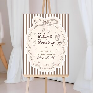 K&ouml;nnte beinhalten: Willkommensschild f&uuml;r eine Babyparty auf einer Staffelei. Das Schild zeigt die Worte "Baby is Brewing" in einem dekorativen Rahmen. Das Schild enth&auml;lt auch den Namen Olivia Smith und das Datum 07.06.202x. Der Hintergrund hat vertikale braune und wei&szlig;e Streifen.