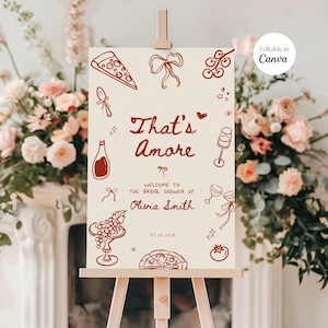 May include: A cream-colored sign on a wooden easel with the text "That's Amore" in red script. The sign welcomes guests to Olivia Smith's bridal shower and features illustrations of pizza, wine, and other Italian-themed icons.