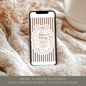 May include: A smartphone displays a baby shower invitation with a decorative frame and the words "Baby is Brewing." The phone has a striped background. The invitation includes details like the baby's name, date, and location. The phone rests on a textured, cream-colored surface.