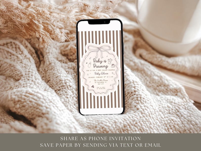May include: A smartphone displays a baby shower invitation with a decorative frame and the words "Baby is Brewing." The phone has a striped background. The invitation includes details like the baby's name, date, and location. The phone rests on a textured, cream-colored surface.