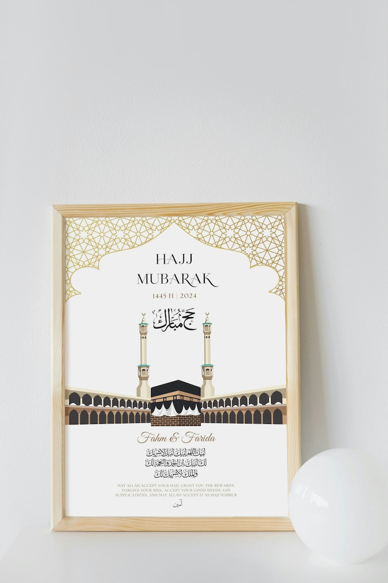 Puede incluir: Una impresi&oacute;n enmarcada con un marco de madera clara. La impresi&oacute;n presenta el texto "HAJJ MUBARAK" y caligraf&iacute;a &aacute;rabe, junto con una ilustraci&oacute;n de la Kaaba y los minaretes. La impresi&oacute;n est&aacute; sobre un fondo blanco.