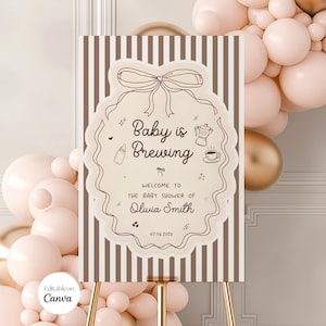 May include: A baby shower sign with the text "Baby is Brewing" in a decorative frame. The sign is on a gold easel, with a striped background. Balloons in shades of pink and gold are in the background.