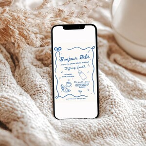 May include: A smartphone displaying a baby shower invitation with the text "Bonjour B&eacute;b&eacute;" and a blue and white illustration of a baby bottle, a cup of coffee, and a croissant. The invitation is for Tiffany Smith and the shower is on Saturday, 12th November at The Smith's House.