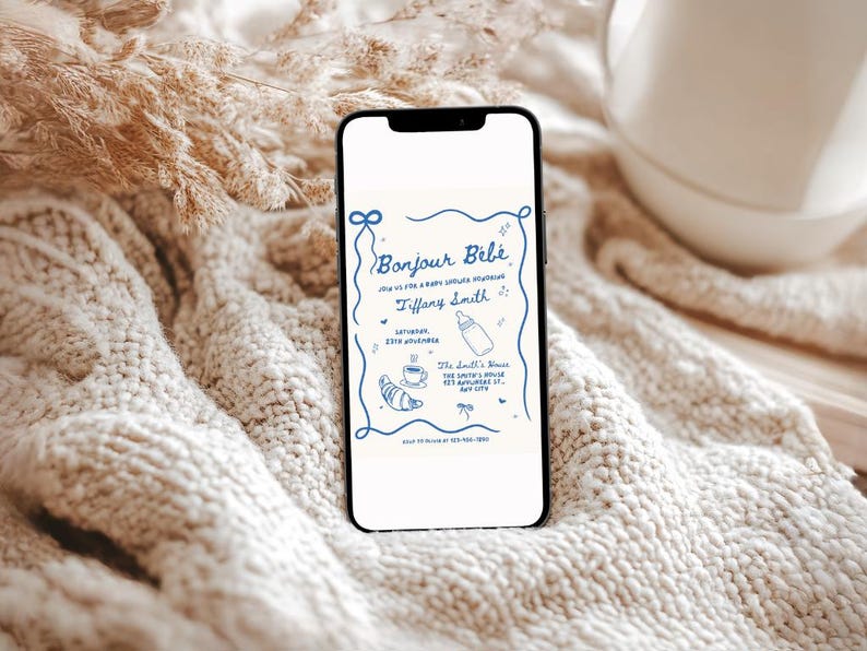 May include: A smartphone displaying a baby shower invitation with the text "Bonjour B&eacute;b&eacute;" and a blue and white illustration of a baby bottle, a cup of coffee, and a croissant. The invitation is for Tiffany Smith and the shower is on Saturday, 12th November at The Smith's House.