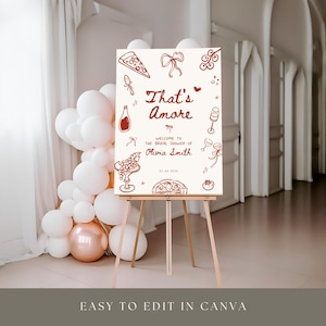 May include: A sign on a wooden easel with the words "That's Amore" in red script, surrounded by illustrations of food and drinks. The sign welcomes guests to a bridal shower. White and rose gold balloons are in the background.