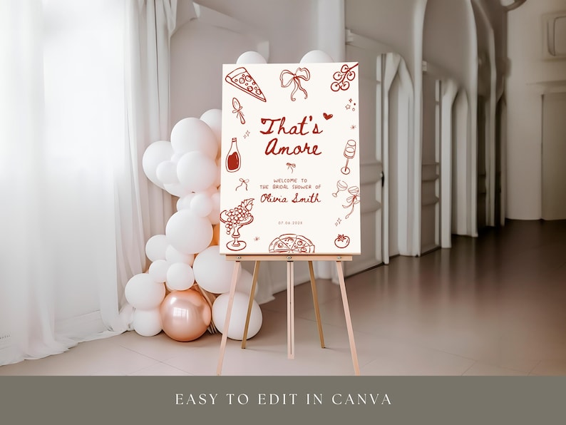 May include: A sign on a wooden easel with the words "That's Amore" in red script, surrounded by illustrations of food and drinks. The sign welcomes guests to a bridal shower. White and rose gold balloons are in the background.