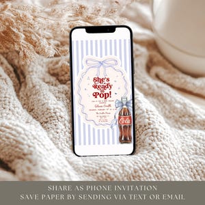 May include: A smartphone displays a digital invitation with the text "She's Ready to Pop!" against a striped background. The invitation features a vintage Coca-Cola bottle illustration with a blue bow. The phone rests on a textured, cream-colored surface.