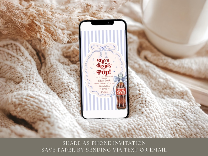 May include: A smartphone displays a digital invitation with the text "She's Ready to Pop!" against a striped background. The invitation features a vintage Coca-Cola bottle illustration with a blue bow. The phone rests on a textured, cream-colored surface.