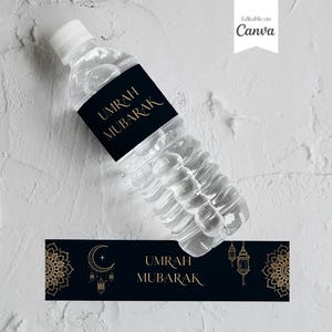 May include: A black and gold water bottle label with the text "Umrah Mubarak". The label has a decorative design with a crescent moon and lanterns.