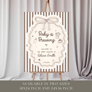 K&ouml;nnte beinhalten: Ein Babyparty-Schild auf einer Staffelei. Das Schild hat einen hellbraunen Hintergrund mit vertikalen Streifen. Ein cremefarbenes Oval in der Mitte zeigt den Text "Baby is Brewing" und "Welcome to the baby shower of Olivia Smith". Dekorative Elemente sind eine Schleife, eine Kaffeekanne und eine Babyflasche.