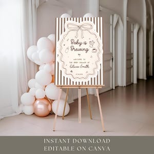 K&ouml;nnte beinhalten: Ein Willkommensschild f&uuml;r eine Babyparty mit dem Text "Baby is Brewing" in einem dekorativen Rahmen. Das Schild steht auf einer h&ouml;lzernen Staffelei, mit Luftballons im Hintergrund. Das Schild ist hellbeige mit braunen Streifen.