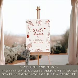 May include: A sign on an easel with the words "That's Amore" in red script, with illustrations of pizza, wine, and grapes. The sign welcomes guests to a bridal shower. The background is a blurred outdoor scene.