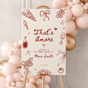 May include: A cream-colored sign on a gold stand with the text "That's Amore" in a handwritten font. The sign welcomes guests to Olivia Smith's bridal shower, with illustrations of pizza, wine, and grapes. A balloon arch is in the background.