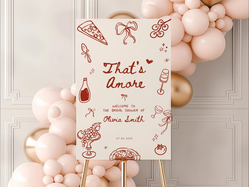 May include: A cream-colored sign on a gold stand with the text "That's Amore" in a handwritten font. The sign welcomes guests to Olivia Smith's bridal shower, with illustrations of pizza, wine, and grapes. A balloon arch is in the background.