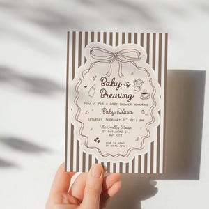 May include: A baby shower invitation with a brown and white striped background. The invitation features the text "Baby is Brewing" in a decorative frame with illustrations of a baby bottle, coffee pot, and coffee cup.