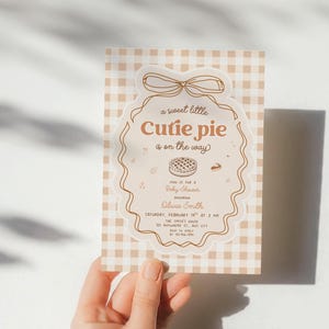 Peut inclure: Une carte d'invitation rectangulaire pour une fête prénatale avec un motif vichy beige et blanc. Le centre présente un cadre festonné avec un nœud, contenant le texte 'a sweet little Cutie pie is on the way' et les détails de l'événement.