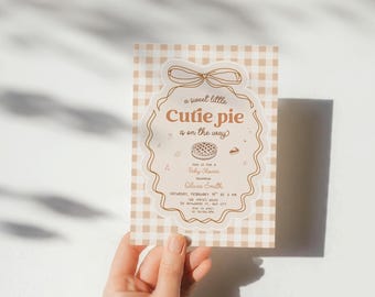 May include: A rectangular baby shower invitation card featuring a beige and white gingham pattern. The center displays a scalloped frame with a bow at the top, containing the text 'a sweet little Cutie pie is on the way' and event details.