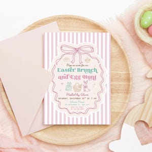 Puede incluir: Una invitaci&oacute;n a un brunch de Pascua y una b&uacute;squeda de huevos con rayas rosas y blancas, un sobre rosa y una bandeja de madera. La invitaci&oacute;n presenta un borde decorativo y texto. Tambi&eacute;n hay un nido con huevos de colores y un conejito de madera.