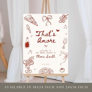 May include: A cream-colored sign on an easel with the phrase "That's Amore" in a handwritten font. The sign features illustrations of pizza, wine bottles, and grapes. Text reads "Welcome to the Bridal Shower of Olivia Smith".