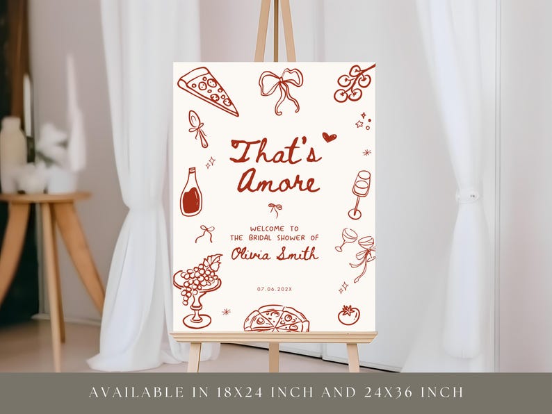 May include: A cream-colored sign on an easel with the phrase "That's Amore" in a handwritten font. The sign features illustrations of pizza, wine bottles, and grapes. Text reads "Welcome to the Bridal Shower of Olivia Smith".