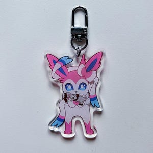 Op de afbeelding: Een roze en blauw anime-personage met lange oren en een strik die twee kleinere roze en blauwe anime-personages vasthoudt. Het personage zit op een transparante acryl sleutelhanger met een zilveren metalen sluiting.
