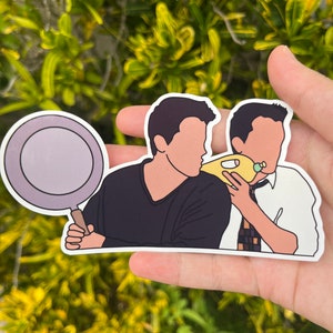 May include: A sticker featuring two cartoon figures from the TV show Friends. One figure holds a frying pan, while the other is eating a sandwich. The sticker has a white border and is set against a green background.