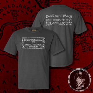 Puede incluir: Una camiseta gris oscuro con un gráfico blanco de un letrero de madera que dice "Motion Picture Location TV Livestock & Equipment Spahn's Ranch". La parte posterior de la camiseta tiene texto blanco que dice "Spahn's Movie Ranch Saddle Horses for Rent 2,50 € días laborables 3,00 € fines de semana Una hora Una hora Festivos".
