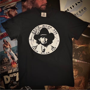 May include: Black t-shirt featuring a white graphic of David Allan Coe in a cowboy hat, encircled by his name. The shirt is a classic crew neck style. The design is distressed for a vintage look.