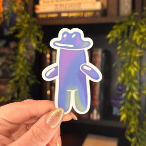 May include: A holographic sticker of a blue, cartoon frog wearing a hat. The frog has a wide smile and is standing with its arms outstretched.