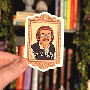 May include: A sticker featuring a cartoon illustration of a man in a framed portrait. The man has brown hair, glasses, and a mustache. The text "Do it lady!" is written below the portrait in a stylized font. The sticker is held by a hand.