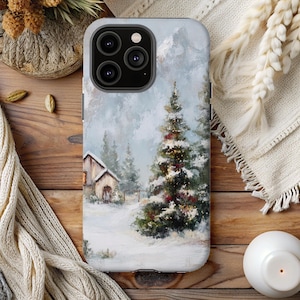 May include: A smartphone case featuring a winter landscape painting with a snow-covered cabin and evergreen trees. The artwork depicts a snowy scene with a Christmas tree, creating a festive and seasonal theme. The case is light gray.