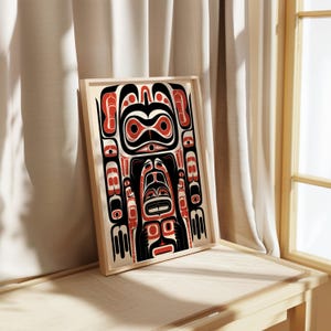 May include: A framed print of a stylized black, red, and white Northwest Coast Indigenous art design. The design features a stylized human figure with a large head and a variety of geometric shapes.