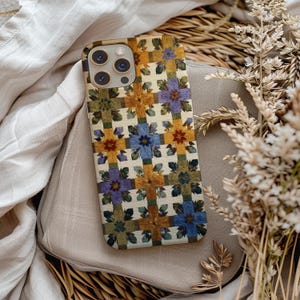 May include: A rectangular smartphone case featuring a repeating grid pattern of pressed flowers in shades of purple, blue, and yellow, set against a cream-coloured background with green foliage accents and a triple-lens camera cutout at the top.