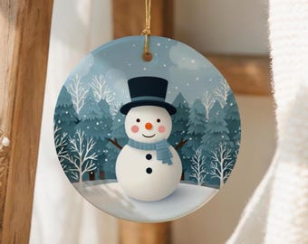 May include: A round Christmas ornament featuring a snowman with a black top hat, scarf, and twig arms. The background shows a snowy forest scene with evergreen trees and a light blue sky. The ornament hangs from a gold string.