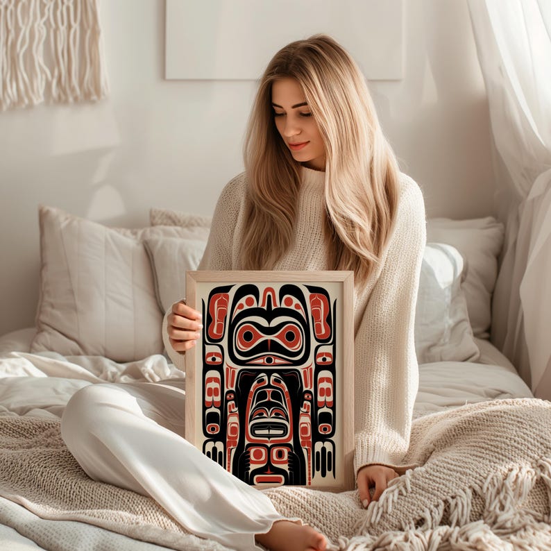 May include: A woman holds a framed print of a colorful Northwest Coast Indigenous art design. The design features a stylized human figure with geometric patterns in red, black, and white.