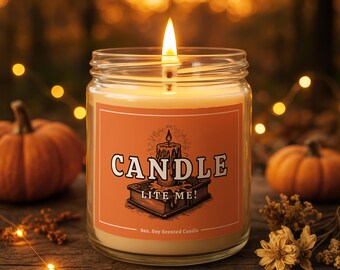 May include: A lit soy-scented candle in a glass jar with an orange label. The label features the word "CANDLE" above the phrase "LITE ME!" and a drawing of a candle on a book. The background includes pumpkins and fairy lights.