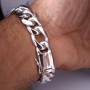 May include: A close-up of a silver curb chain bracelet. The bracelet is made of thick, interlocking links and features a secure clasp. The polished metal reflects light, highlighting the bracelet's design. The bracelet is worn on a wrist.
