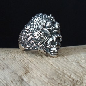 Op de afbeelding: Gedetailleerde zilveren ring met een versierd schedelontwerp. De ring heeft ingewikkelde gravures en een getextureerde afwerking. De schedel is het centrale kenmerk, tegen een houten achtergrond. Het ontwerp is complex.
