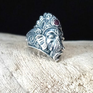 May include: A silver ring with an intricate design of the Hindu deity Ganesha. The ring features a red gemstone in the center of Ganesha's forehead.