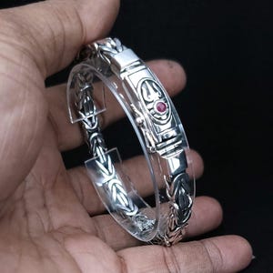Könnte beinhalten: Ein silbernes Armband mit einem detaillierten Design und einem roten Edelstein im Verschluss.
