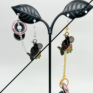 May include: A pair of dangle earrings featuring a black crow with red and green floral accents. One earring has a silver chain and a pink donut charm, while the other has a gold chain and a pink donut charm.