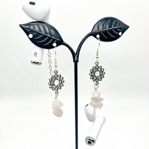 May include: A pair of silver earrings with a geometric design and pink crystal accents hanging from a black earring stand.